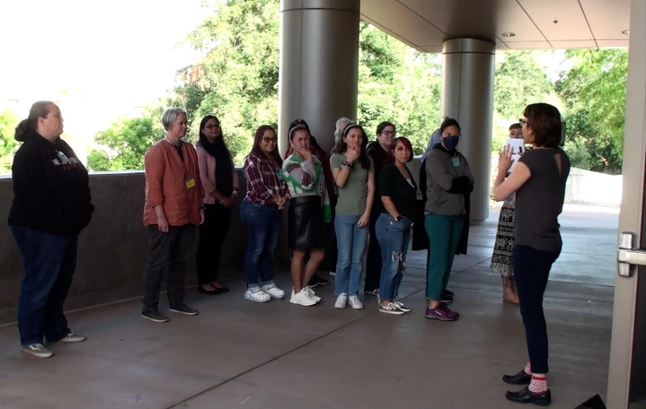 A group of 12 adults are standing outside while a facilitator speaks to them