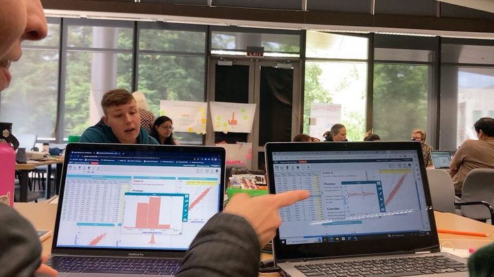 Two computer screens shows data points represented on graphs as two adults at a table discuss the data on the screen 