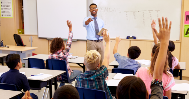 Math teacher stands at the front of the room while elementary students raise their hands at desks